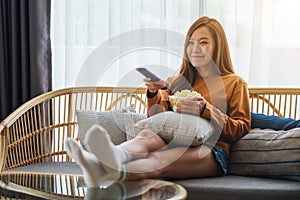 A beautiful young woman eating popcorn and searching channel with remote control to watch tv while sitting on