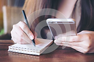 Closeup image of a business woman writing on notebook while using and holding mobile phone on wooden table background