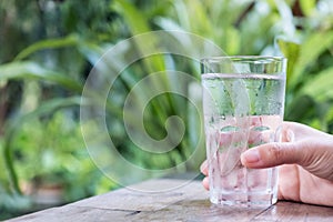 Closeup image of a hand holding a glass of cold water on wooden table with green nature