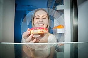 Closeup image of hungry young woman taking high-caloric donut from refrigerator at night. Concept of unhealthy nutrition