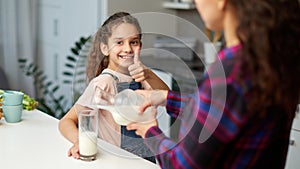 Closeup image of a mom pours her cute smiling daughter milk for breakfast, looking at camera shows thumb.