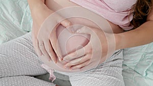 Closeup image of pregnant woman stroking belly and making shape of heart. Concept of expecting child, love and parenting