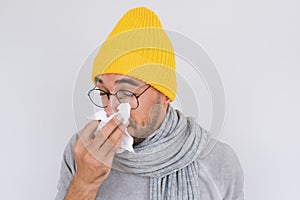 Closeup image of sick handsome man wearing grey sweater, yellow hat and glasses, blowing nose and sneeze into tissue. Male have