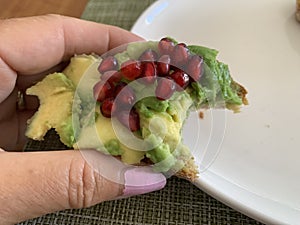 Closeup image of a woman holding a half eaten slice of sourdough avocado toast topped with pomegranate arils