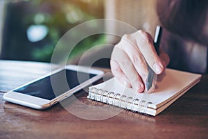 Closeup image of a woman holding pencil and writing on notebook with white mobile phone on wooden table