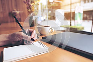 Closeup image of woman`s hand writing on a blank notebook with laptop , tablet and coffee cup on wooden table