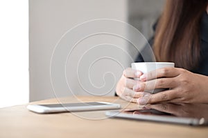 A woman`s hands holding a cup of coffee with smart phone and tablet pc on wooden table in cafe