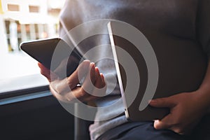 A woman using and looking at a black smart phone while holding laptop computer