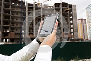Closeup image of young man in white shirt holding digital tablet against building under construction. You can insert