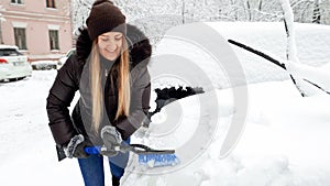 Closeup image of young smiling blond woman in brown coat and jeans trying to clean up snow covered car by brush after a