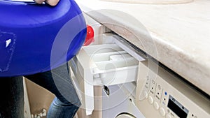 Closeup image of young woman pouring liquid detergent in washing machine from big bottle