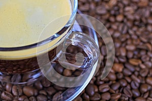 Closeup of isoalted transparent glass cup, saucer and handle with black coffee whitecap crema, blurred roasted coffee beans