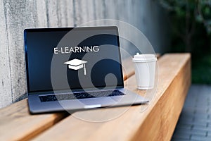 Closeup laptop with inscription on screen e-learning and image of square academic cap and cup of coffee on bench. Online education