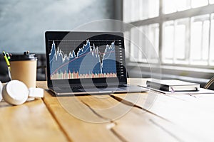 Closeup of laptop with stock market analyst monitor and cups, headphones, coffee on a table in a cafe.