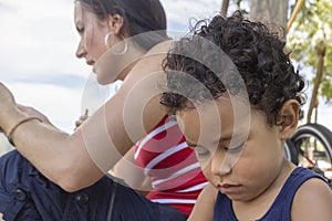 Closeup of a little boy sitting next to his mother with a sad face looking down