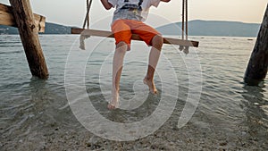 Closeup of little boy swinging on the sea beach and touching ocean waves with feet. Holiday, summer vacation and tourism
