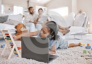 Closeup of a little cute girl using a laptop and wireless headphones while laying on the floor in the lounge. Hispanic