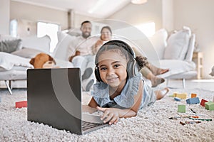 Closeup of a little cute girl using a laptop and wireless headphones while laying on the floor in the lounge. Hispanic