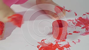 Closeup of a little girl with her hands dirty in red paint draws her fingers on a large sheet of white paper sitting on