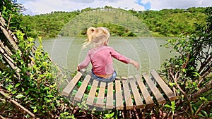 Closeup Little Girl Sits on Bench In Clipped Bush Watches Lake