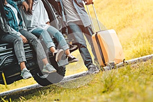 Closeup lower body of group of friends relaxing on SUV car trunk with trolly luggage along road trip with autumn mountain hill