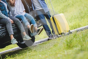 Closeup lower body of group of friends relaxing on SUV car trunk with trolly luggage along road trip with autumn mountain hill