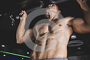 Closeup of a macho shirtless man in his late 20s hangs on a pull up bar. Training lats and back. Doing a set of pullups or muscle