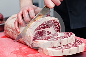 Closeup macro cook`s hand cut ribeye marbled beef steak with knife on red plastic cutting board on metal table in restaurant