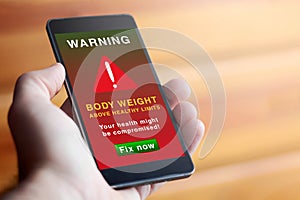 A closeup of male hand holding modern mobile phone with red screen and warning symbol with text warning about overweight. Weight