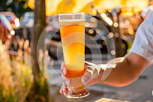 Closeup of male hand holding up one glass of beer in sunny day in summer. Blur orange umbrella background