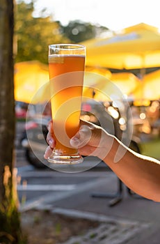 Closeup of male hand holding up one glass of beer in sunny day in summer. Blur orange umbrella background