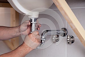 Closeup male plumber worker in blue denim uniform, overalls, fixing sink in bathroom with tile wall. Professional plumbing repair