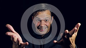 Closeup of a man in black clothes on black background. 4k. Slow motion. man beckons his finger to himself, smiles and