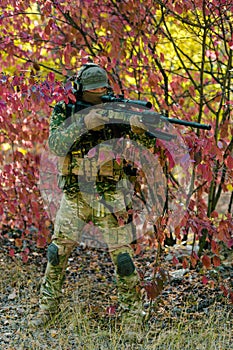 Closeup man in green uniform with sniper rifle. Airsoft soldier stand in red bush. Vertical photo photo