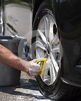 Closeup man hand washing new car alloy silver wheels with big scrub brush, soap suds and bucket