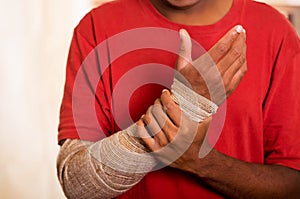 Closeup man in red shirt wearing large grey bandage over lower right arm, supporting with other hand