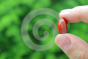 Closeup Man`s Fingers Holding a Red Softgel Supplement Pill Against Blurry Green Foliage