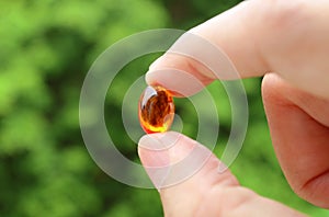 Closeup Man`s Fingers Holding a Transparent Supplement Gelcap Against Blurry Green Foliage