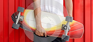 Closeup man`s hand holding the surfskate board in hip position in front of red galvanized steel sheet wall