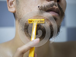 Closeup man use yellow shaver shaving messy beard and mustache on his face in bathroom