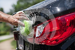 Closeup man washing new black car rear tail light with big soft sponge, soap suds