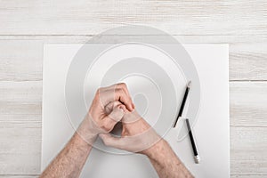 Closeup masculine folded hands on wooden panel with white paper and broken black pencil in top view.