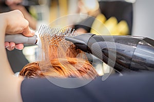 Closeup of master's hand with blow-drying and hairbrush blowing female red hair in a salon.