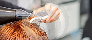 Closeup of master& x27;s hand with blow-drying and hairbrush blowing female red hair in a salon.