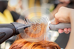 Closeup of master's hand with blow-drying and hairbrush blowing female red hair in a salon.