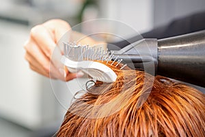 Closeup of master's hand with blow-drying and hairbrush blowing female red hair in a salon.