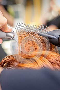 Closeup of master's hand with blow-drying and hairbrush blowing female red hair in a salon.