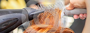 Closeup of master's hand with blow-drying and hairbrush blowing female red hair in a salon.