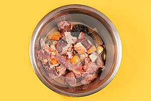 Closeup metal pet bowl with chicken necks, beef meat and bones, paunch, raw egg and carrot pieces.