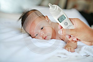 Closeup mother hand takes temperature for infant baby boy with high pressure of ear thermometer on bed at home